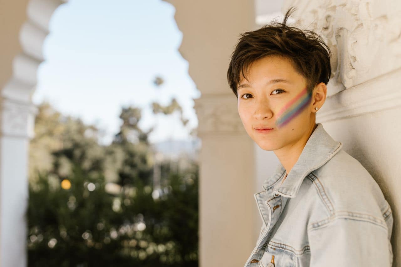 Portrait of a person with short hair and rainbow face paint in a sunlit outdoor location.